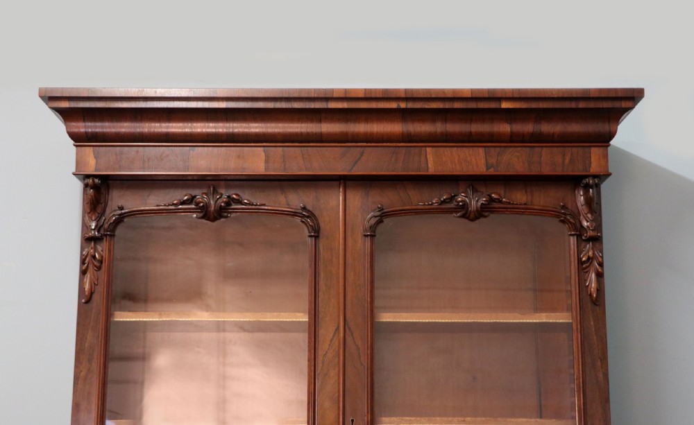 An early Victorian glazed secretaire bookcase cabinet in dark figured wood with carved arch details, a central desk drawer, and lower cupboard doors. View VIII