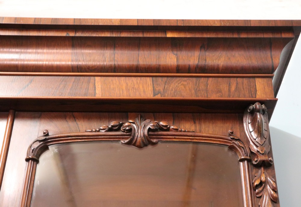 An early Victorian glazed secretaire bookcase cabinet in dark figured wood with carved arch details, a central desk drawer, and lower cupboard doors. View X