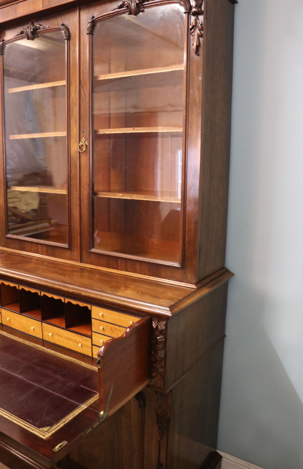 An early Victorian glazed secretaire bookcase cabinet in dark figured wood with carved arch details, a central desk drawer, and lower cupboard doors. View XIX
