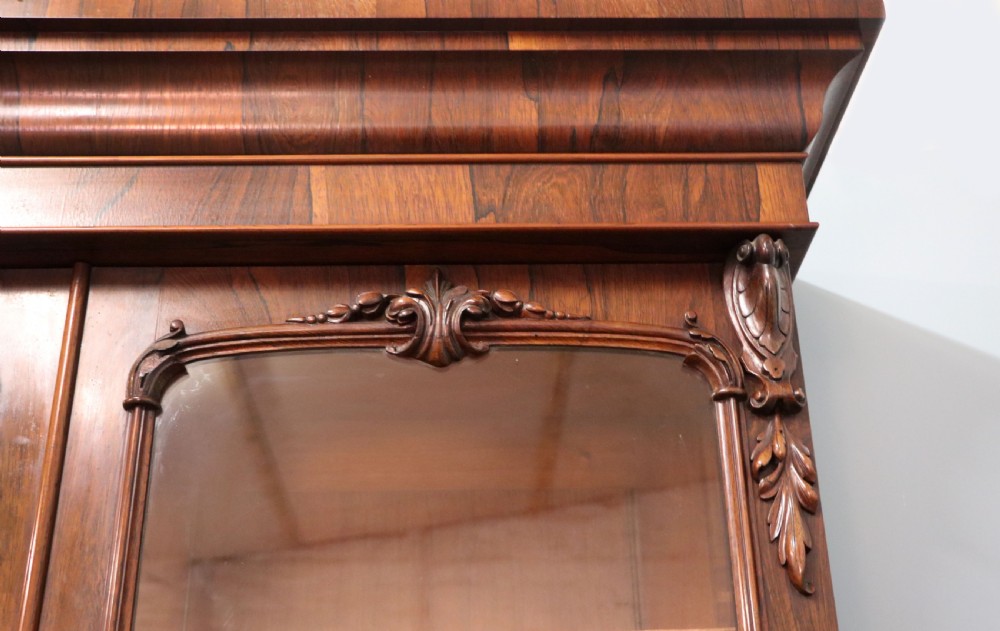 An early Victorian glazed secretaire bookcase cabinet in dark figured wood with carved arch details, a central desk drawer, and lower cupboard doors. View XX