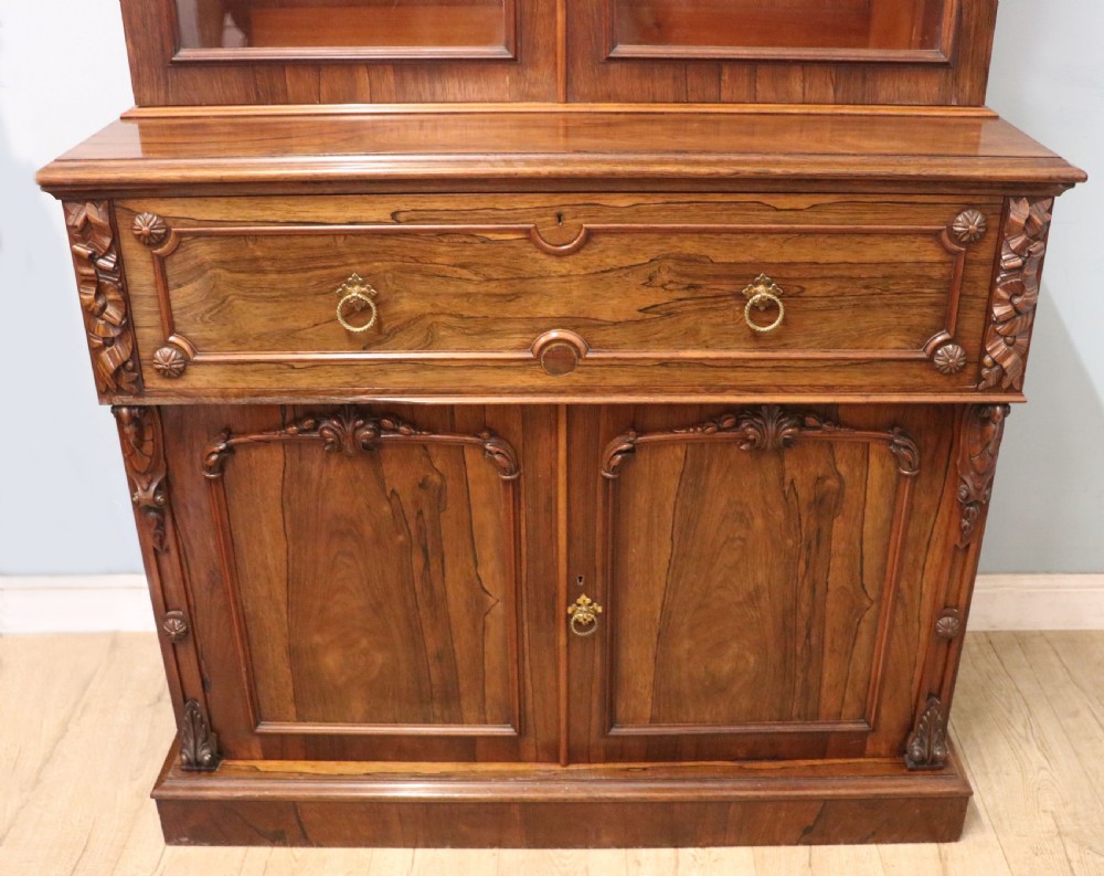 An early Victorian glazed secretaire bookcase cabinet in dark figured wood with carved arch details, a central desk drawer, and lower cupboard doors. View XXIX