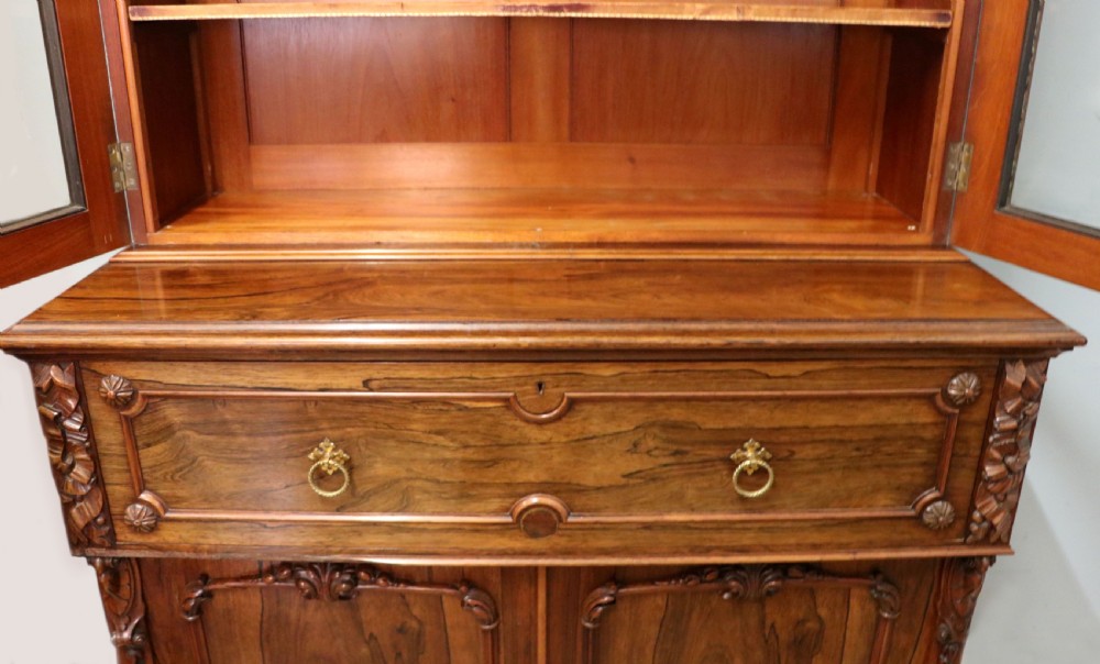 An early Victorian glazed secretaire bookcase cabinet in dark figured wood with carved arch details, a central desk drawer, and lower cupboard doors. View XXX
