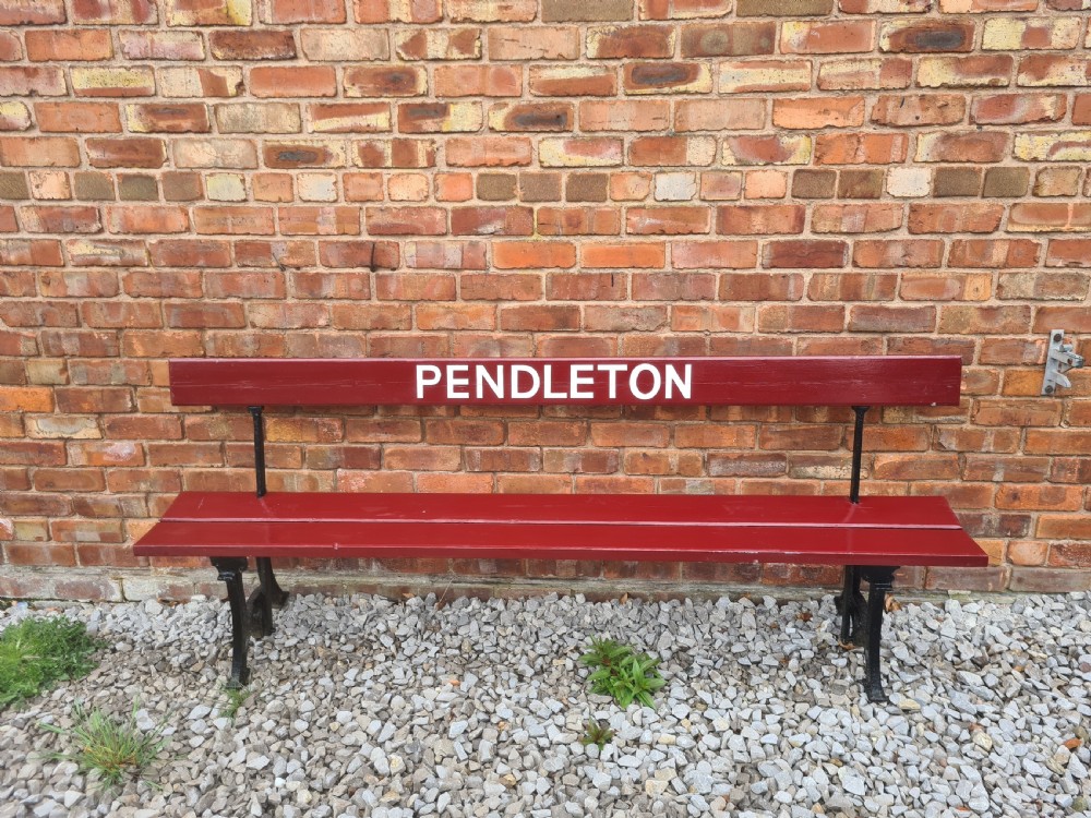 A late nineteenth-century railway station bench with deep red painted wooden slats displaying white stencilled lettering that reads PENDLETON, supported on black cast iron legs.