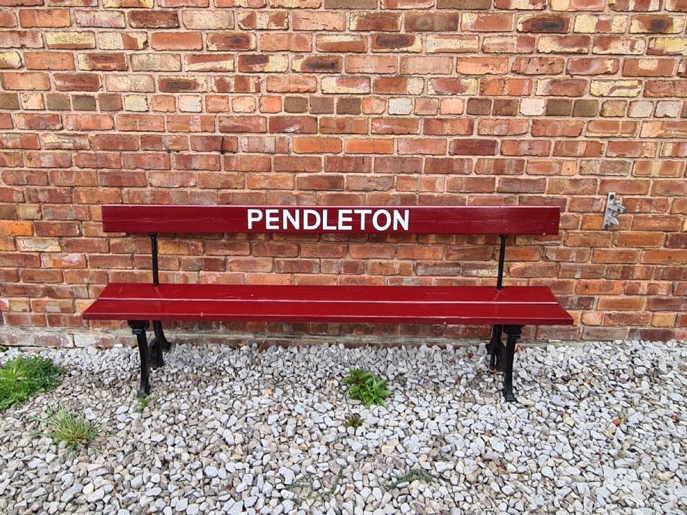 A late nineteenth-century railway station bench with deep red painted wooden slats displaying white stencilled lettering that reads PENDLETON, supported on black cast iron legs. View II