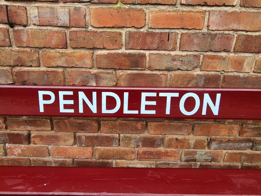 A late nineteenth-century railway station bench with deep red painted wooden slats displaying white stencilled lettering that reads PENDLETON, supported on black cast iron legs. View III