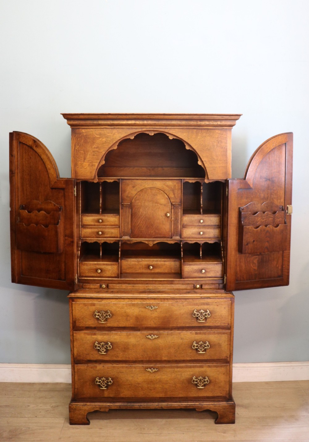 A Georgian style warm brown oak bureau cabinet featuring two upper arched doors opening to a fitted interior of pigeonholes and small drawers above three long drawers with brass handles.