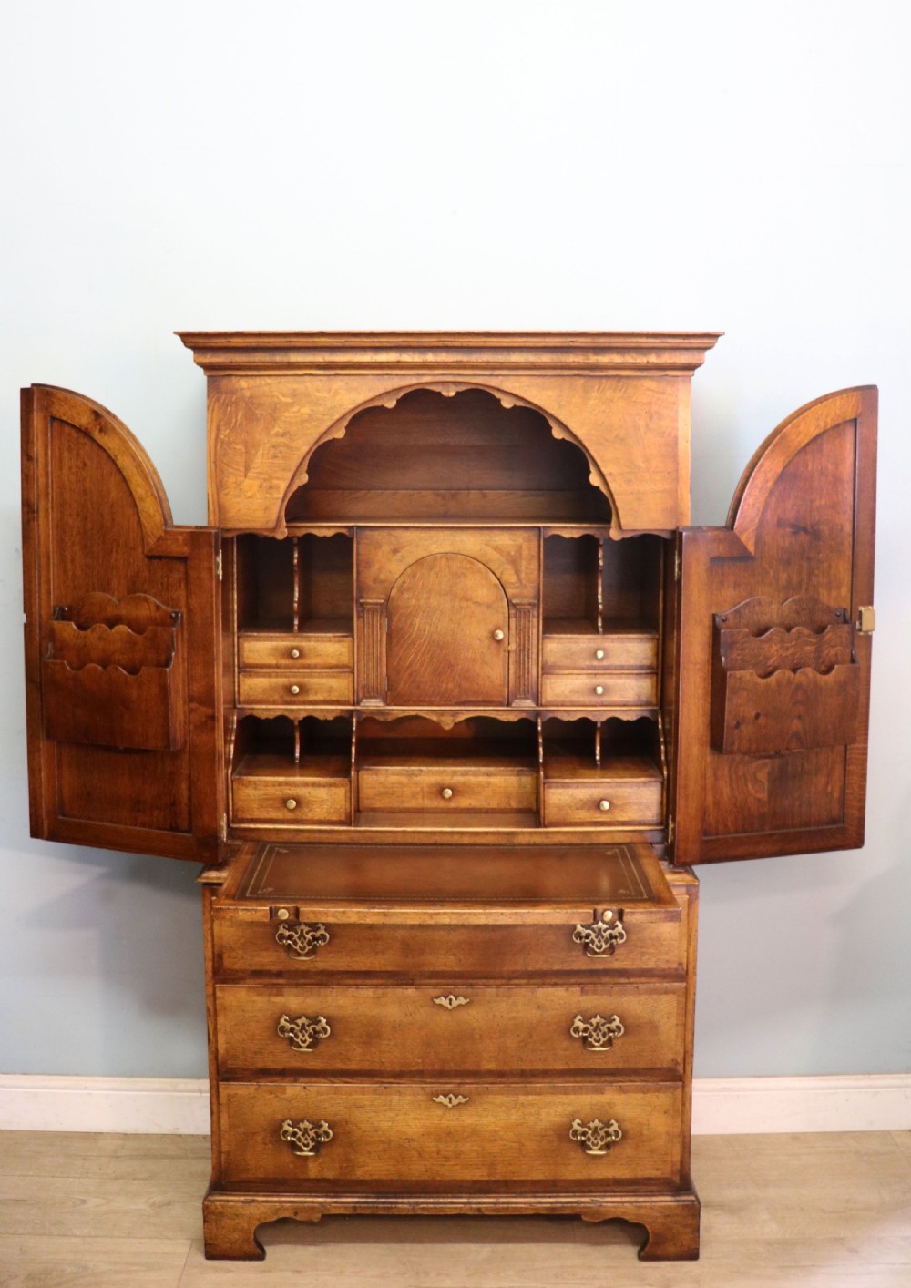 A Georgian style warm brown oak bureau cabinet featuring two upper arched doors opening to a fitted interior of pigeonholes and small drawers above three long drawers with brass handles. View II
