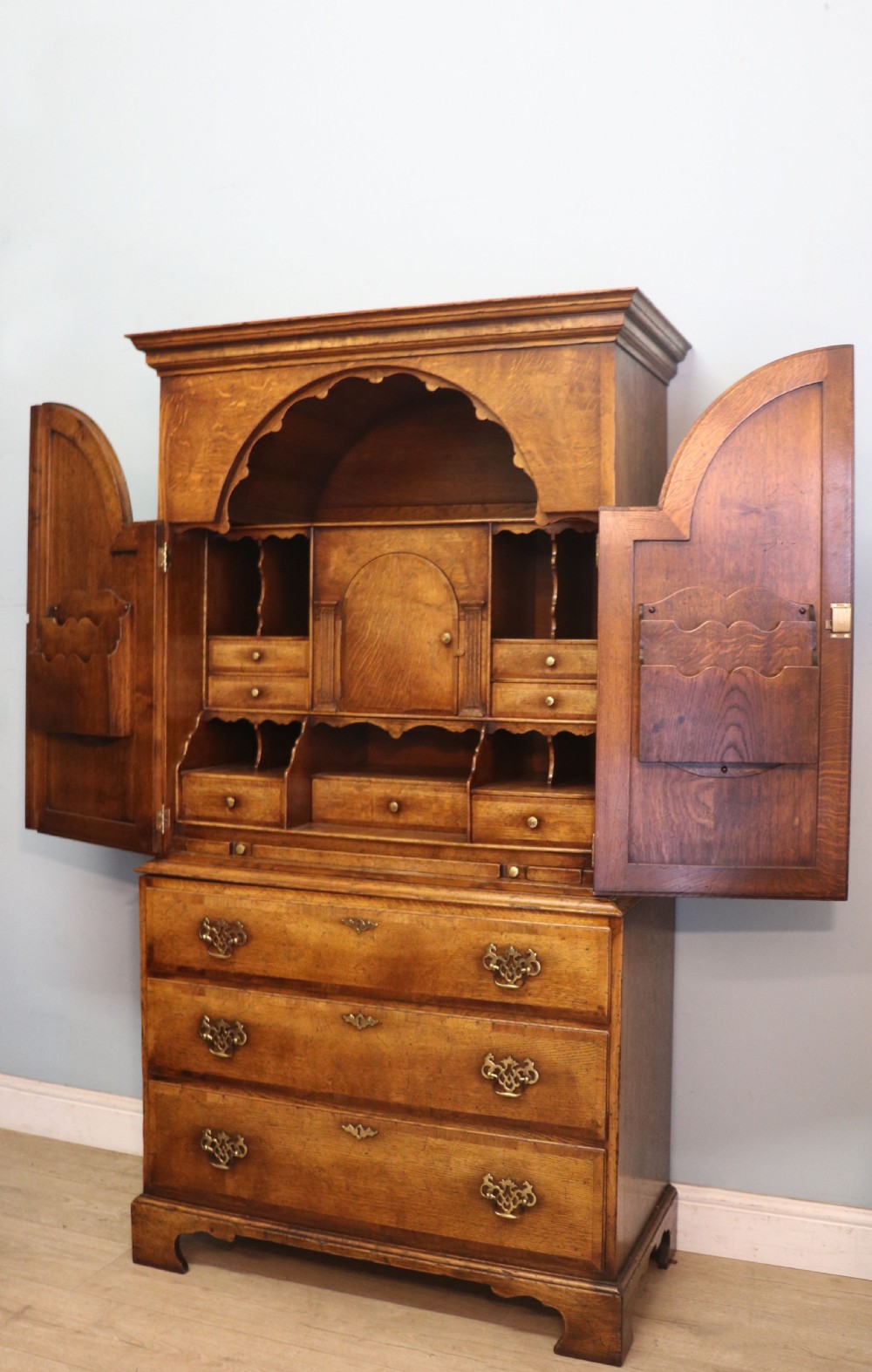 A Georgian style warm brown oak bureau cabinet featuring two upper arched doors opening to a fitted interior of pigeonholes and small drawers above three long drawers with brass handles. View V
