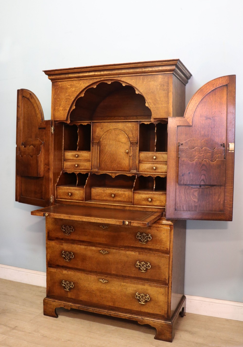 A Georgian style warm brown oak bureau cabinet featuring two upper arched doors opening to a fitted interior of pigeonholes and small drawers above three long drawers with brass handles. View IX