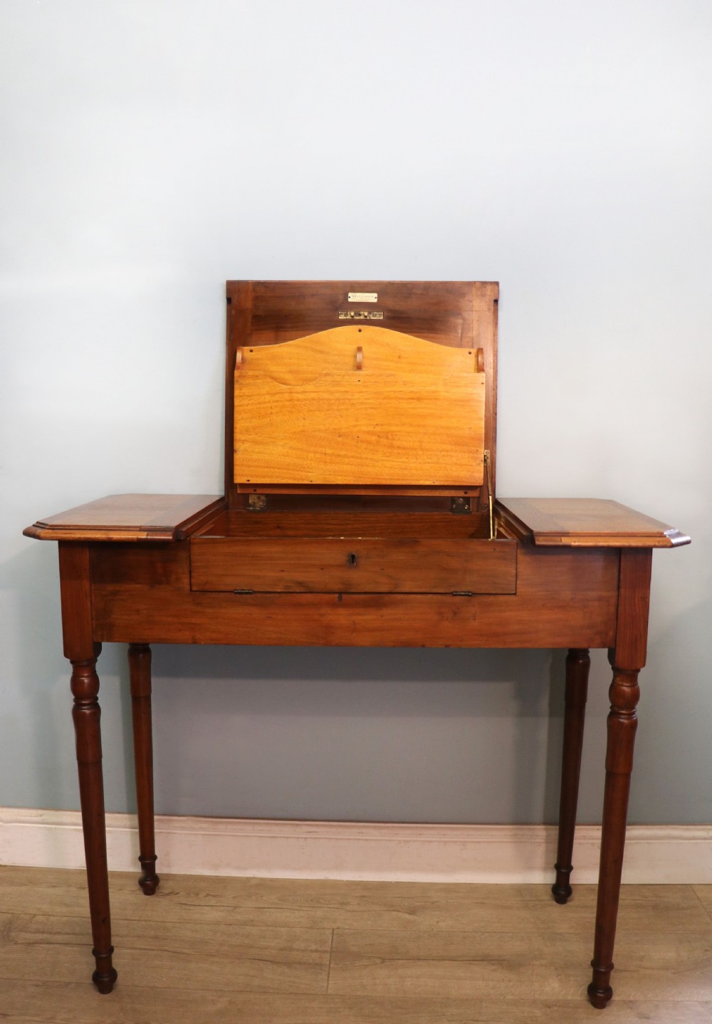 A brown walnut metamorphic writing desk featuring turned legs, side flaps, and an open top section exposing internal stationery compartments.