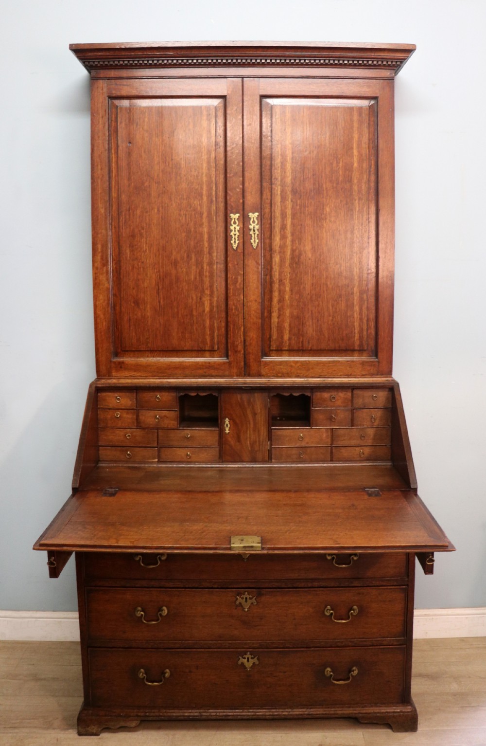 A George III oak bureau bookcase featuring a dentil cornice above two panelled upper doors, a slant-front writing slope opening to a fitted interior, and three long drawers below with brass handles.