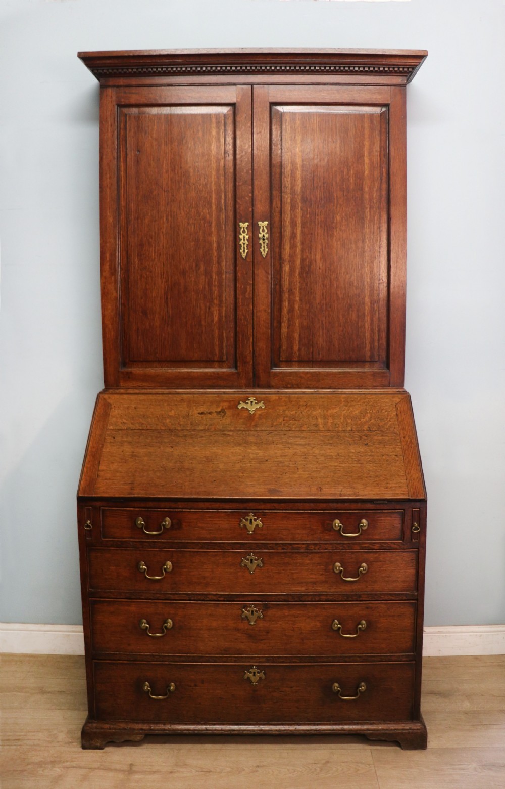 A George III oak bureau bookcase featuring a dentil cornice above two panelled upper doors, a slant-front writing slope opening to a fitted interior, and three long drawers below with brass handles. View II