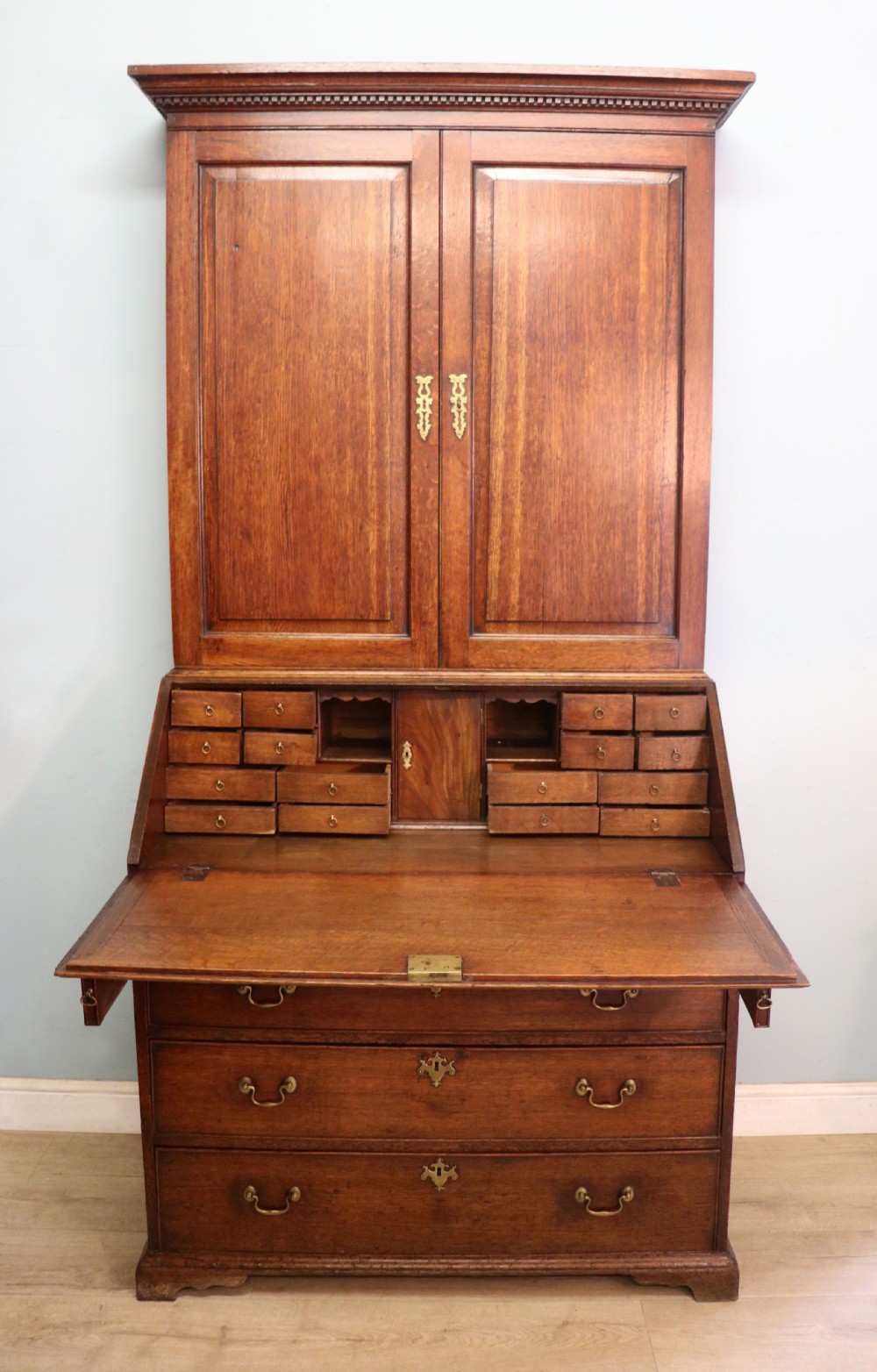A George III oak bureau bookcase featuring a dentil cornice above two panelled upper doors, a slant-front writing slope opening to a fitted interior, and three long drawers below with brass handles. View III
