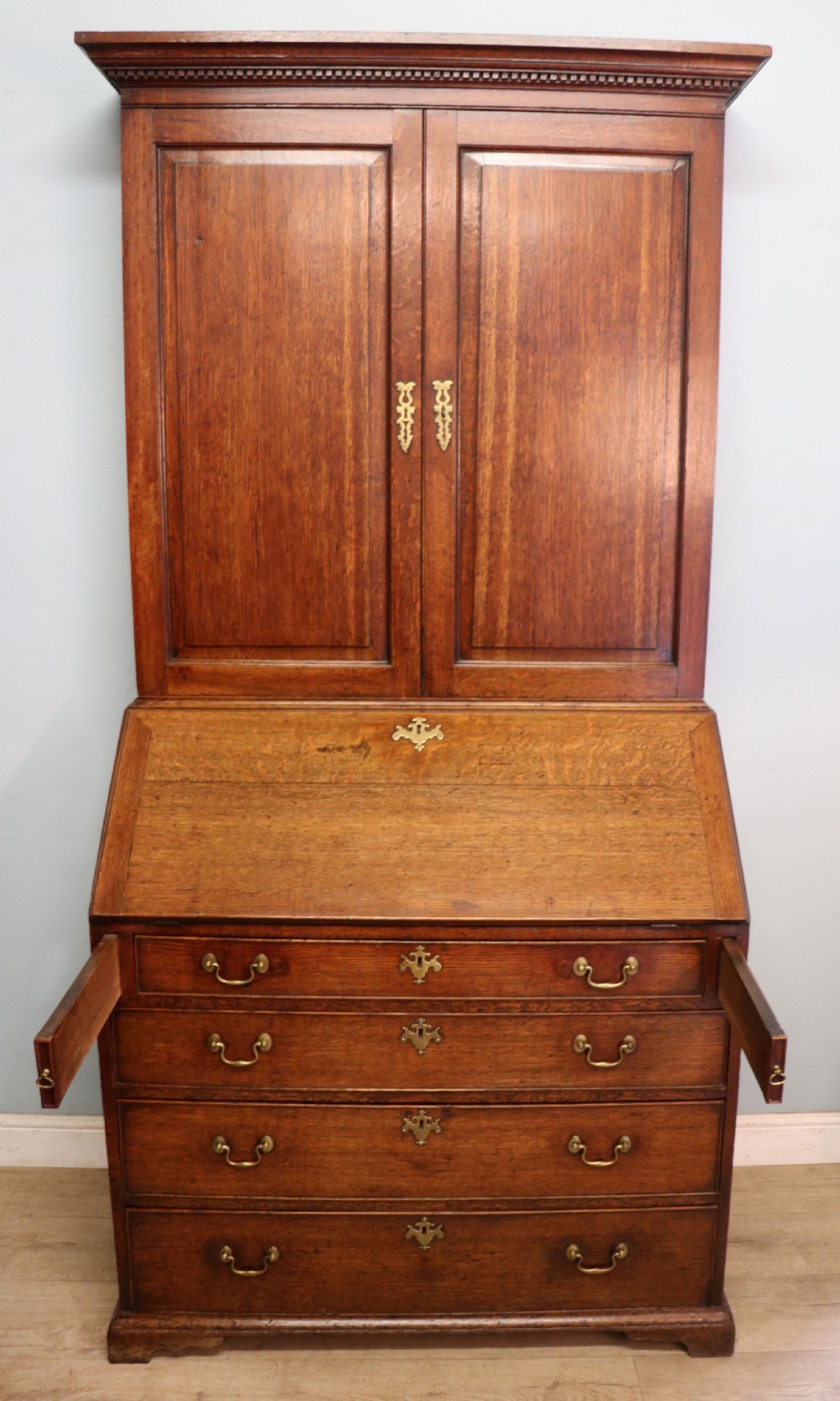 A George III oak bureau bookcase featuring a dentil cornice above two panelled upper doors, a slant-front writing slope opening to a fitted interior, and three long drawers below with brass handles. View IV