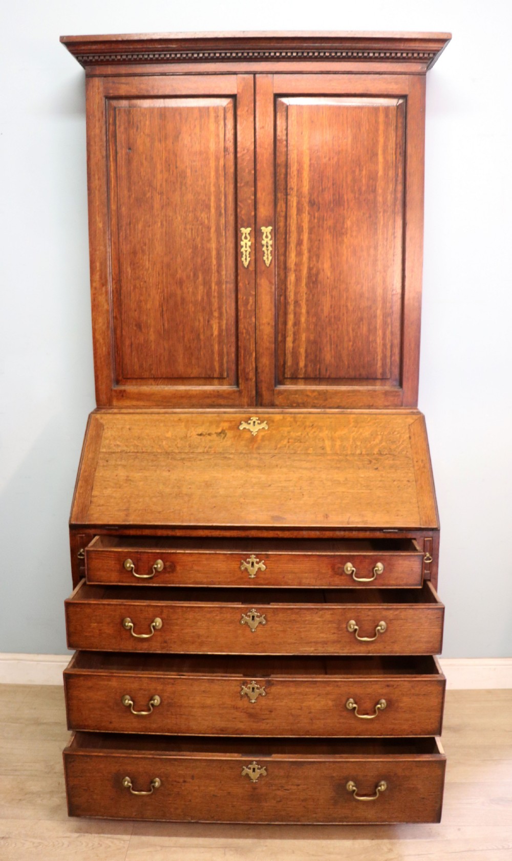 A George III oak bureau bookcase featuring a dentil cornice above two panelled upper doors, a slant-front writing slope opening to a fitted interior, and three long drawers below with brass handles. View V