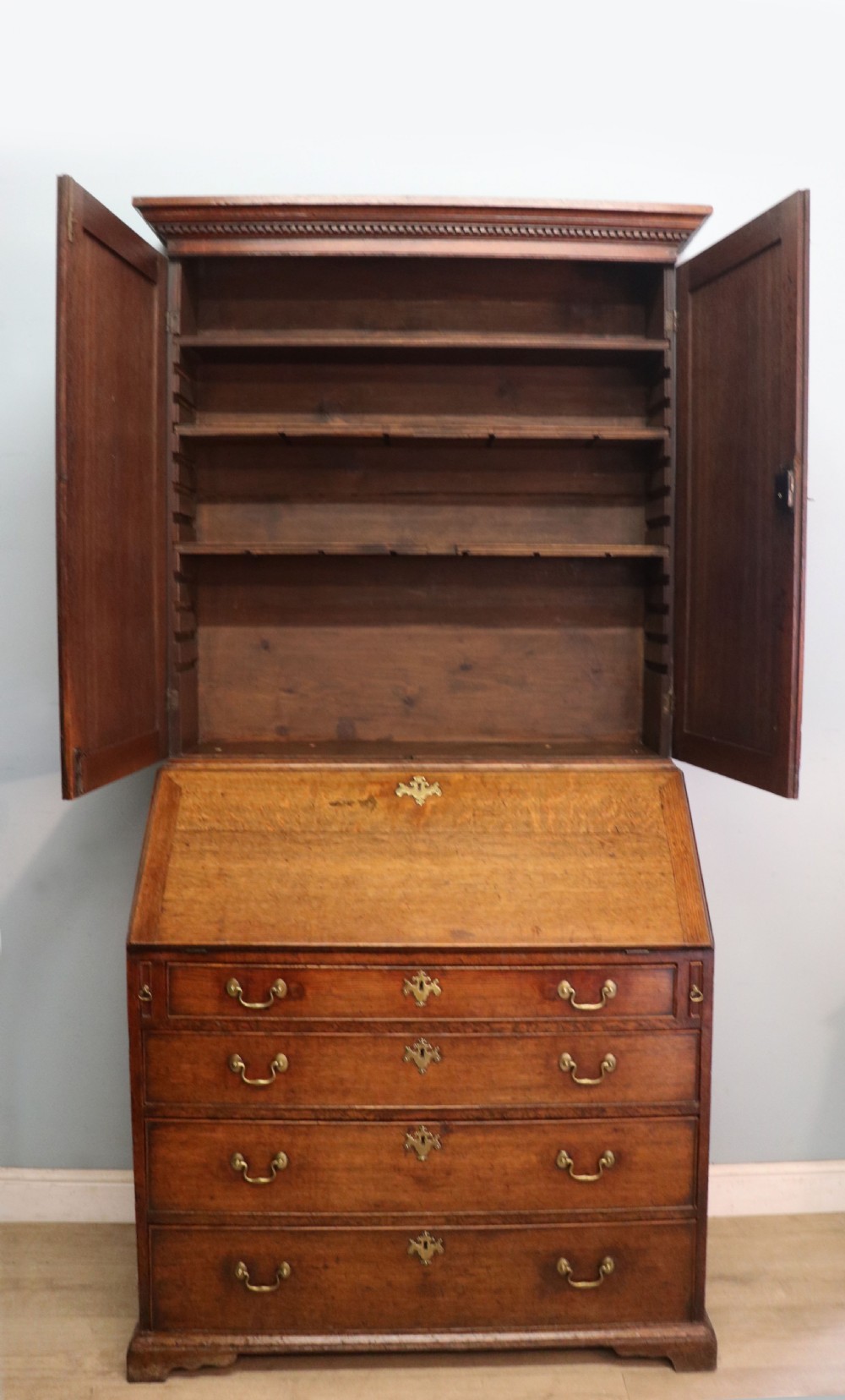 A George III oak bureau bookcase featuring a dentil cornice above two panelled upper doors, a slant-front writing slope opening to a fitted interior, and three long drawers below with brass handles. View VI