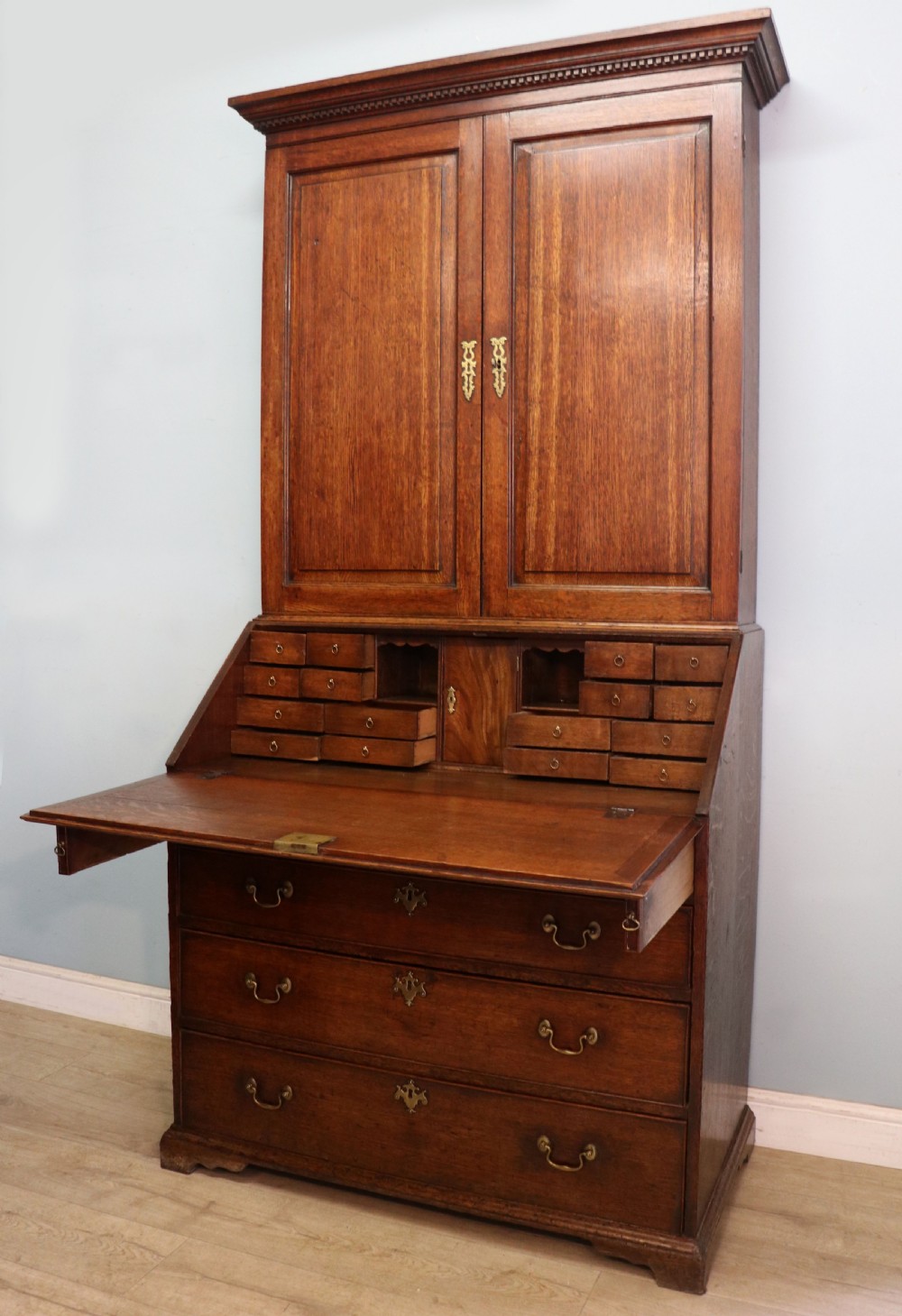 A George III oak bureau bookcase featuring a dentil cornice above two panelled upper doors, a slant-front writing slope opening to a fitted interior, and three long drawers below with brass handles. View VII