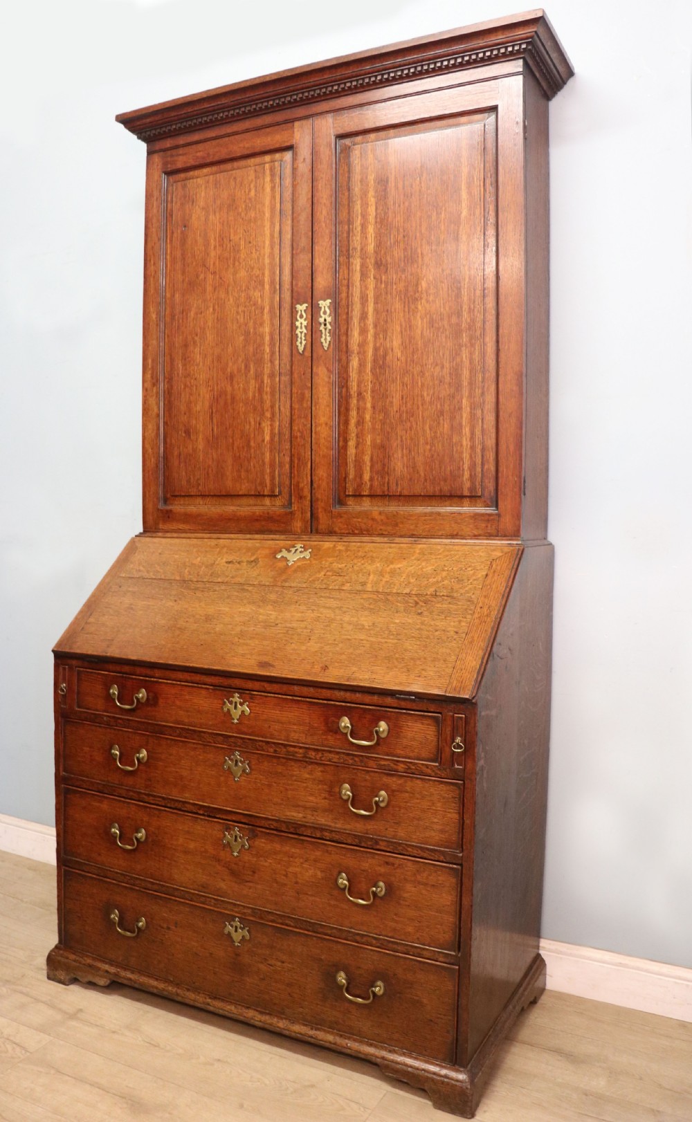 A George III oak bureau bookcase featuring a dentil cornice above two panelled upper doors, a slant-front writing slope opening to a fitted interior, and three long drawers below with brass handles. View VIII