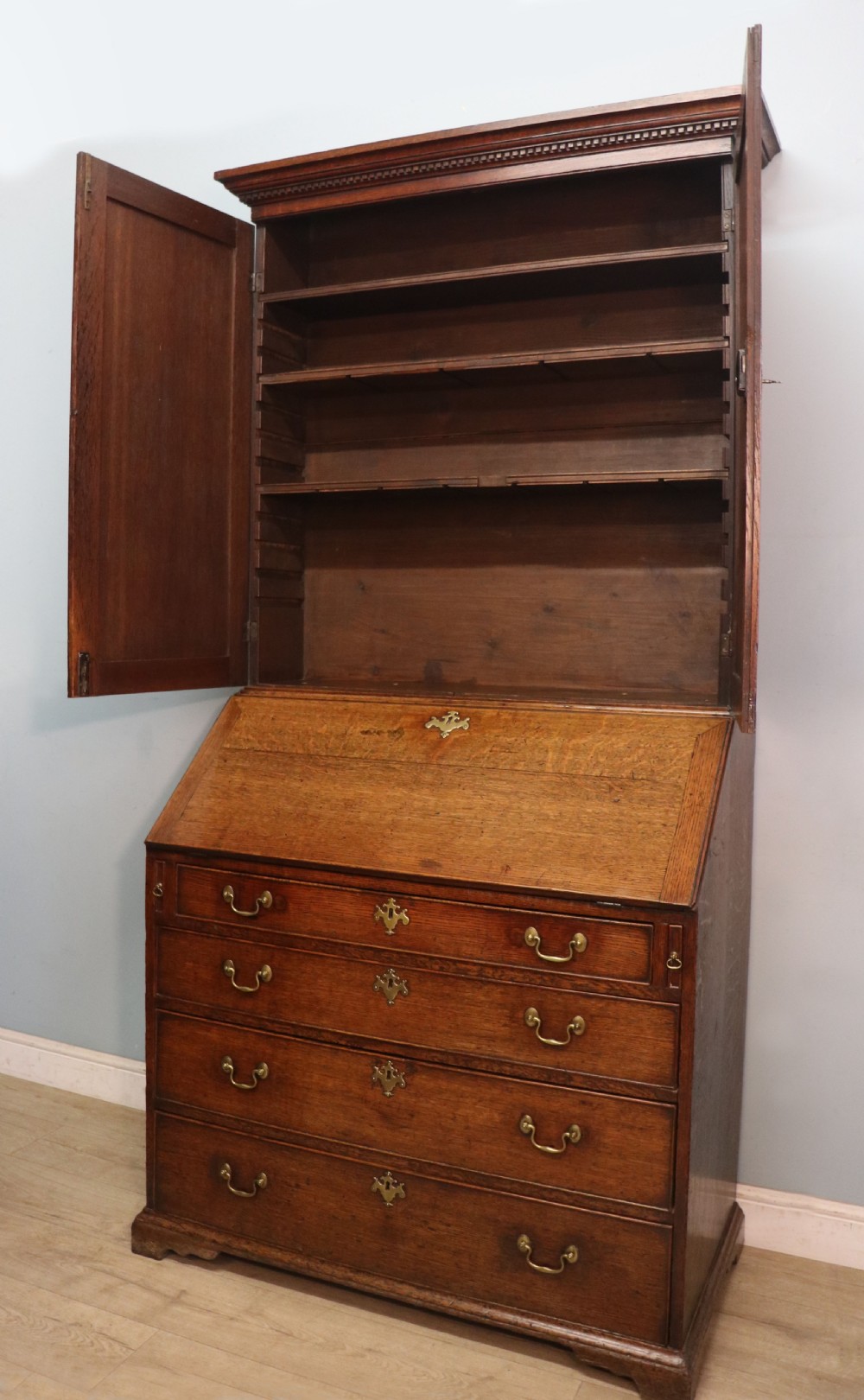 A George III oak bureau bookcase featuring a dentil cornice above two panelled upper doors, a slant-front writing slope opening to a fitted interior, and three long drawers below with brass handles. View IX