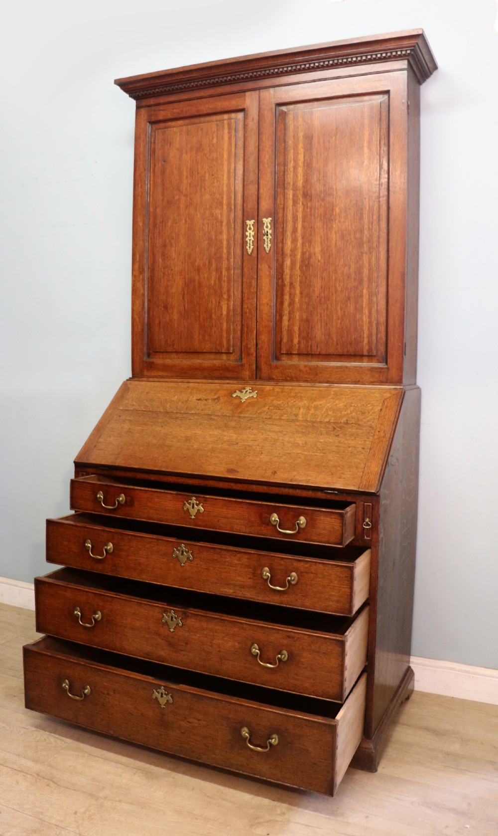 A George III oak bureau bookcase featuring a dentil cornice above two panelled upper doors, a slant-front writing slope opening to a fitted interior, and three long drawers below with brass handles. View X