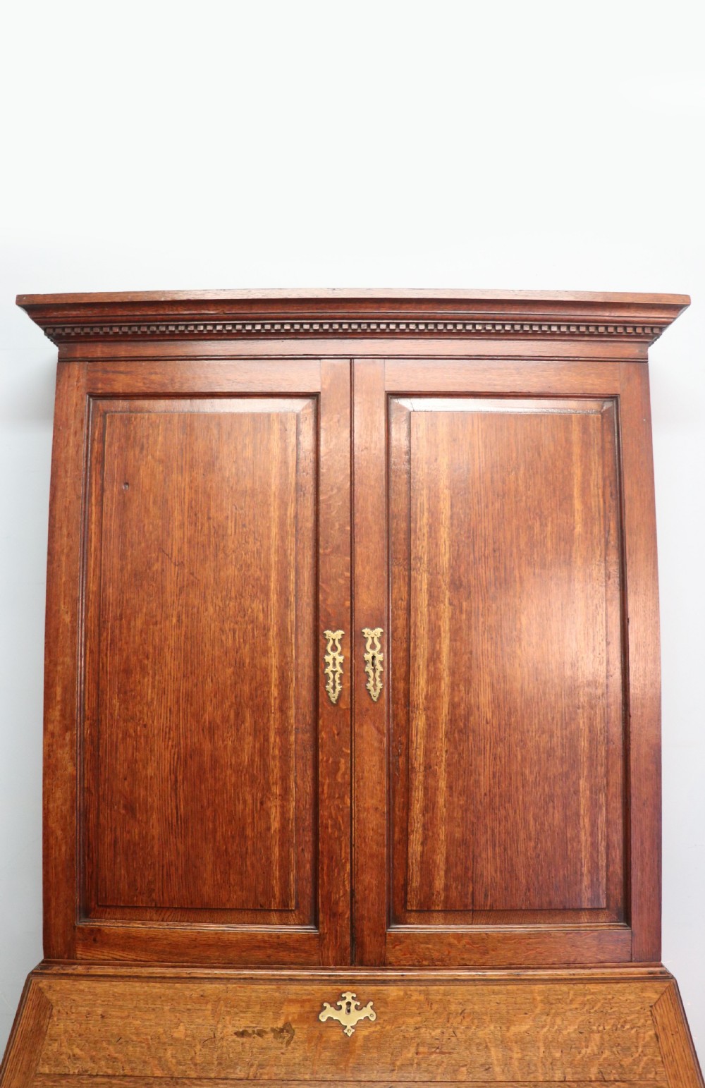 A George III oak bureau bookcase featuring a dentil cornice above two panelled upper doors, a slant-front writing slope opening to a fitted interior, and three long drawers below with brass handles. View XI