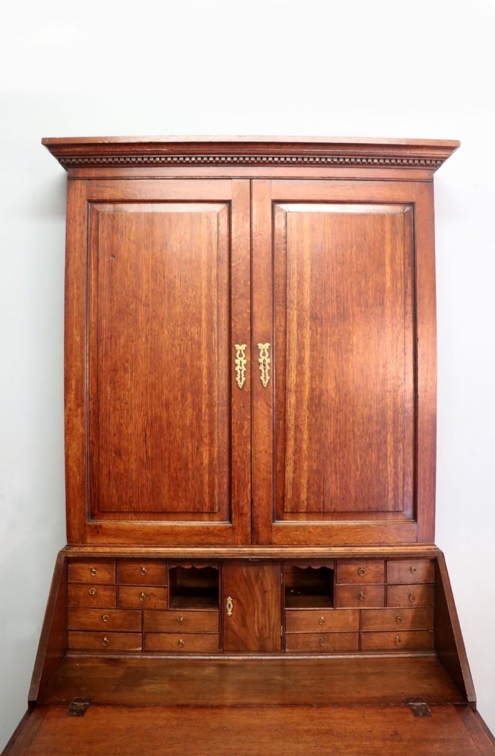 A George III oak bureau bookcase featuring a dentil cornice above two panelled upper doors, a slant-front writing slope opening to a fitted interior, and three long drawers below with brass handles. View XII