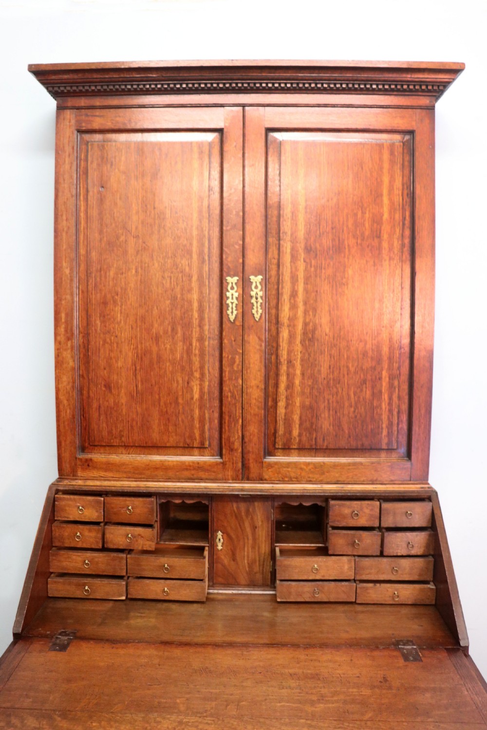 A George III oak bureau bookcase featuring a dentil cornice above two panelled upper doors, a slant-front writing slope opening to a fitted interior, and three long drawers below with brass handles. View XIII