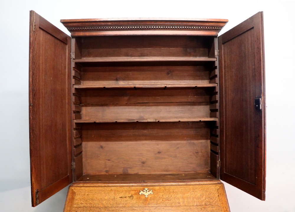 A George III oak bureau bookcase featuring a dentil cornice above two panelled upper doors, a slant-front writing slope opening to a fitted interior, and three long drawers below with brass handles. View XVI