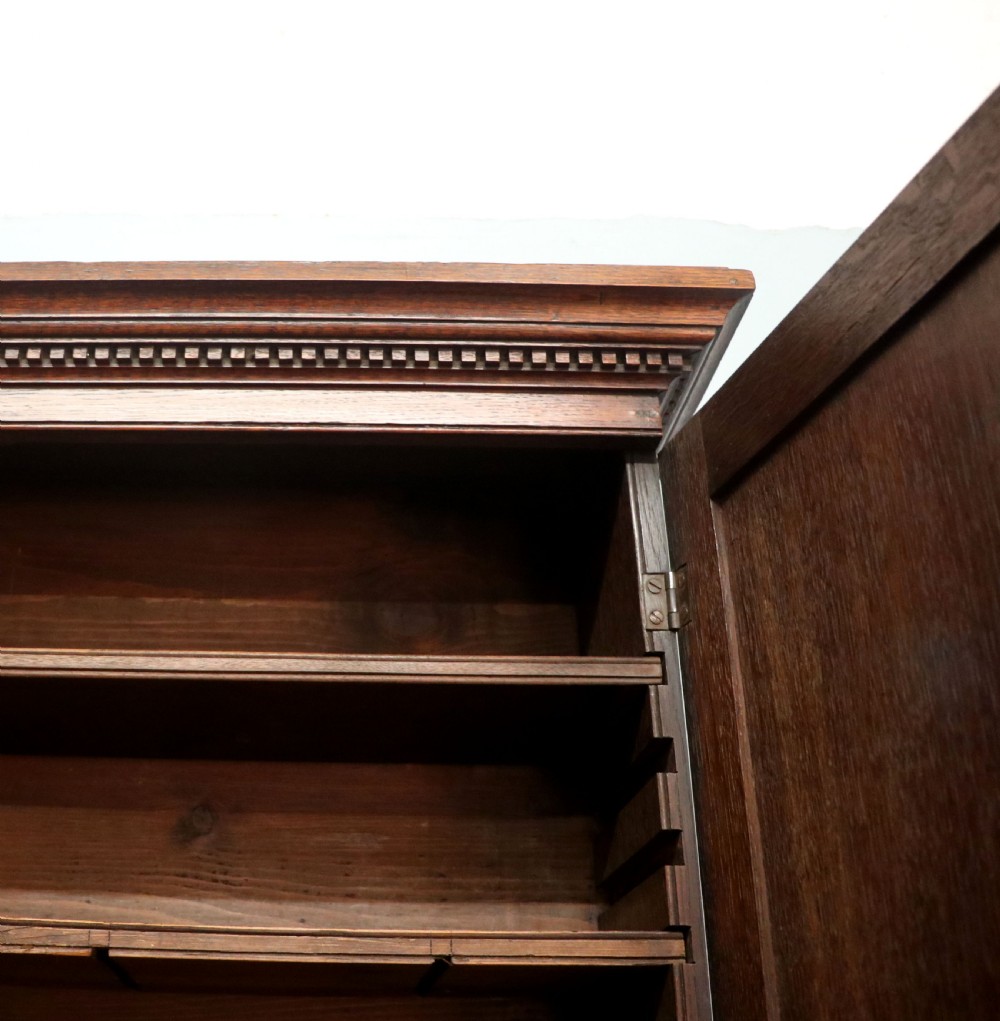 A George III oak bureau bookcase featuring a dentil cornice above two panelled upper doors, a slant-front writing slope opening to a fitted interior, and three long drawers below with brass handles. View XVII