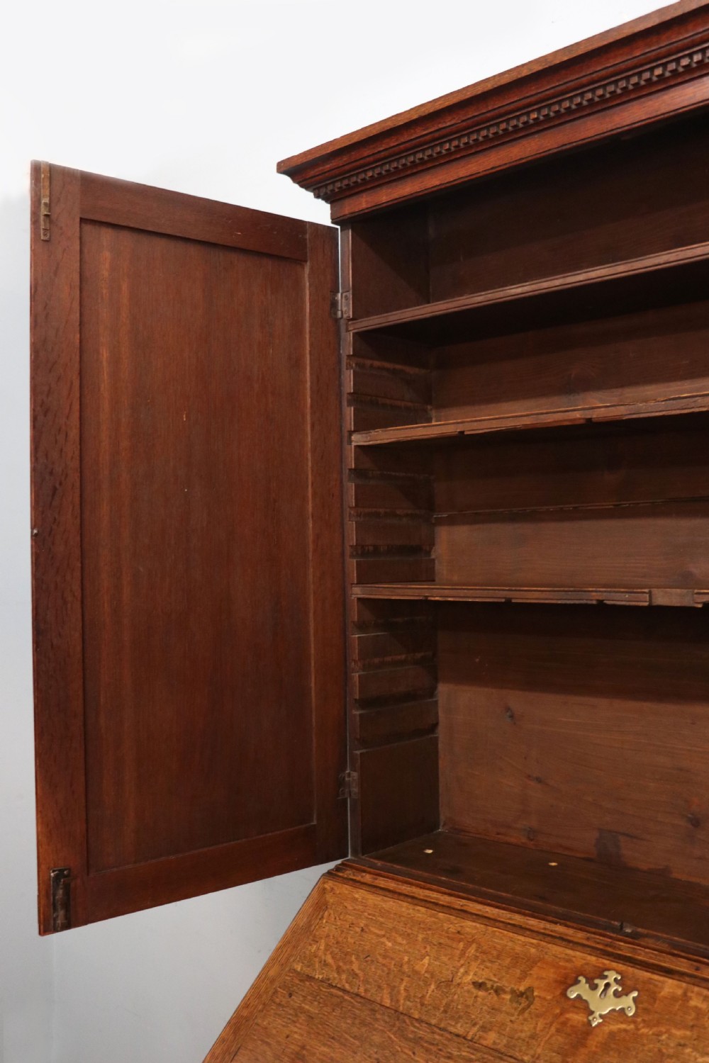 A George III oak bureau bookcase featuring a dentil cornice above two panelled upper doors, a slant-front writing slope opening to a fitted interior, and three long drawers below with brass handles. View XVIII