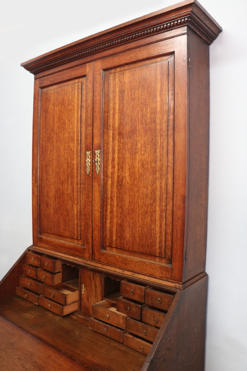 A George III oak bureau bookcase featuring a dentil cornice above two panelled upper doors, a slant-front writing slope opening to a fitted interior, and three long drawers below with brass handles. View XXIII