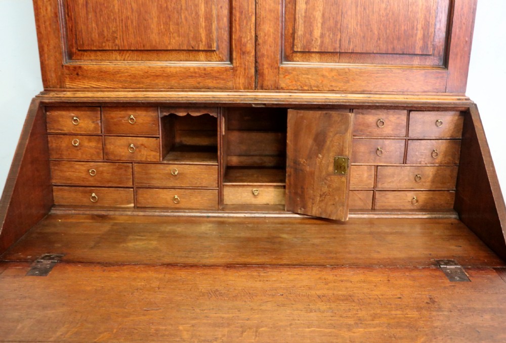 A George III oak bureau bookcase featuring a dentil cornice above two panelled upper doors, a slant-front writing slope opening to a fitted interior, and three long drawers below with brass handles. View XXVII