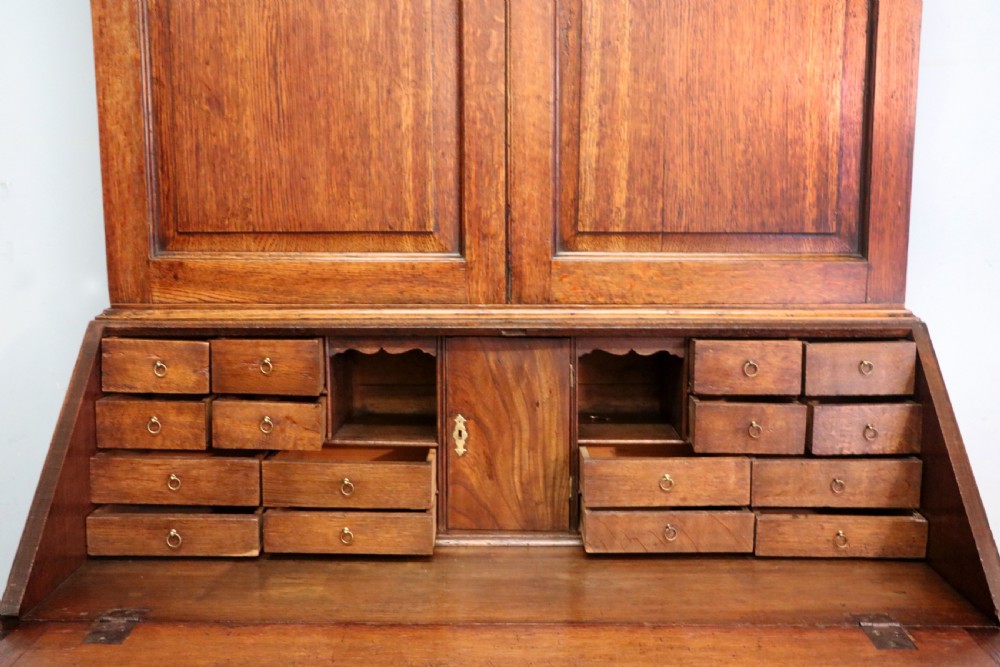 A George III oak bureau bookcase featuring a dentil cornice above two panelled upper doors, a slant-front writing slope opening to a fitted interior, and three long drawers below with brass handles. View XXVIII