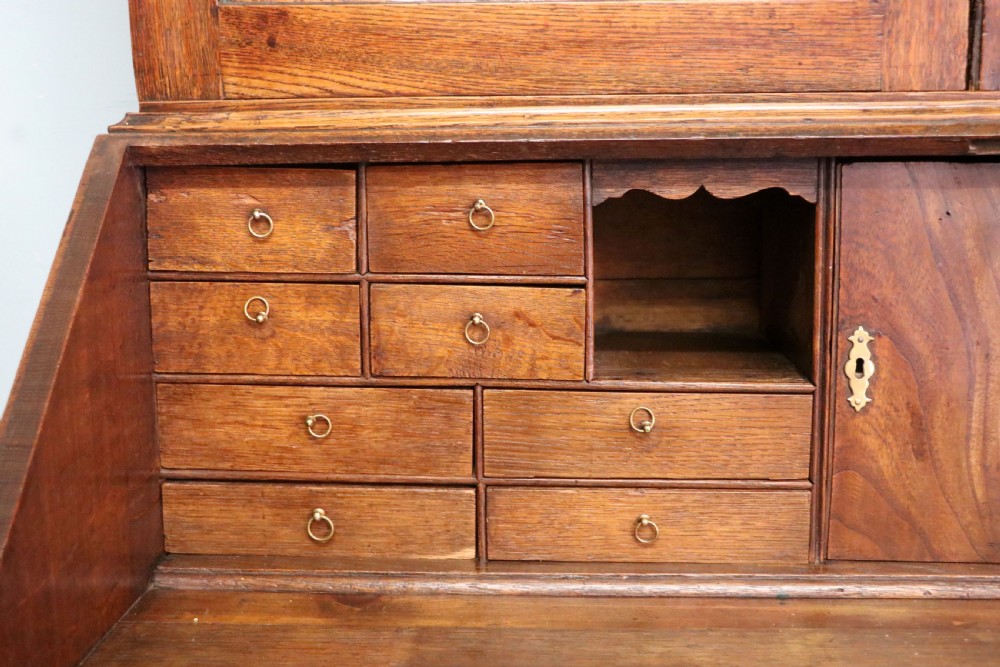 A George III oak bureau bookcase featuring a dentil cornice above two panelled upper doors, a slant-front writing slope opening to a fitted interior, and three long drawers below with brass handles. View XXIX