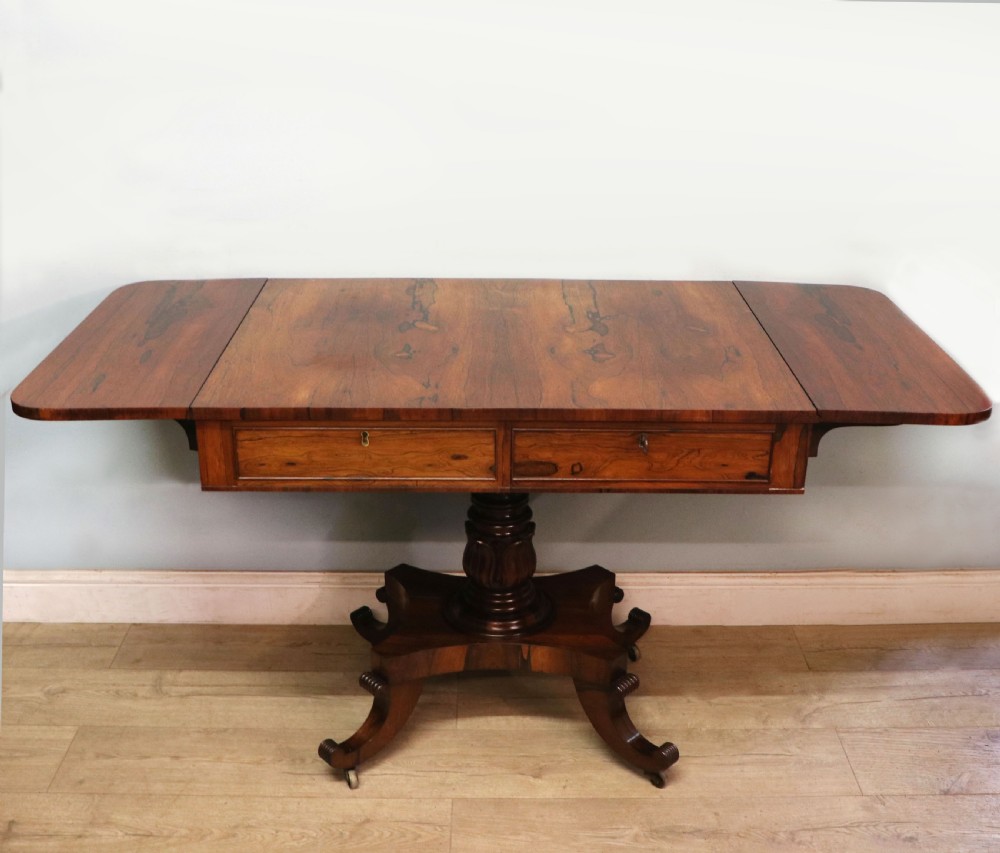 A late Georgian drop-leaf sofa table crafted from dark figured rosewood with a turned pedestal stem, two frieze drawers, and splayed quadruped legs with brass castors.