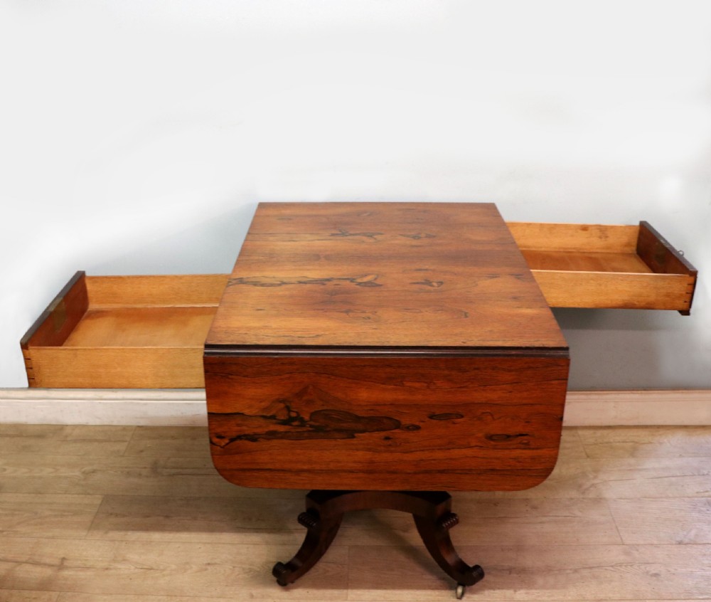 A late Georgian drop-leaf sofa table crafted from dark figured rosewood with a turned pedestal stem, two frieze drawers, and splayed quadruped legs with brass castors. View V