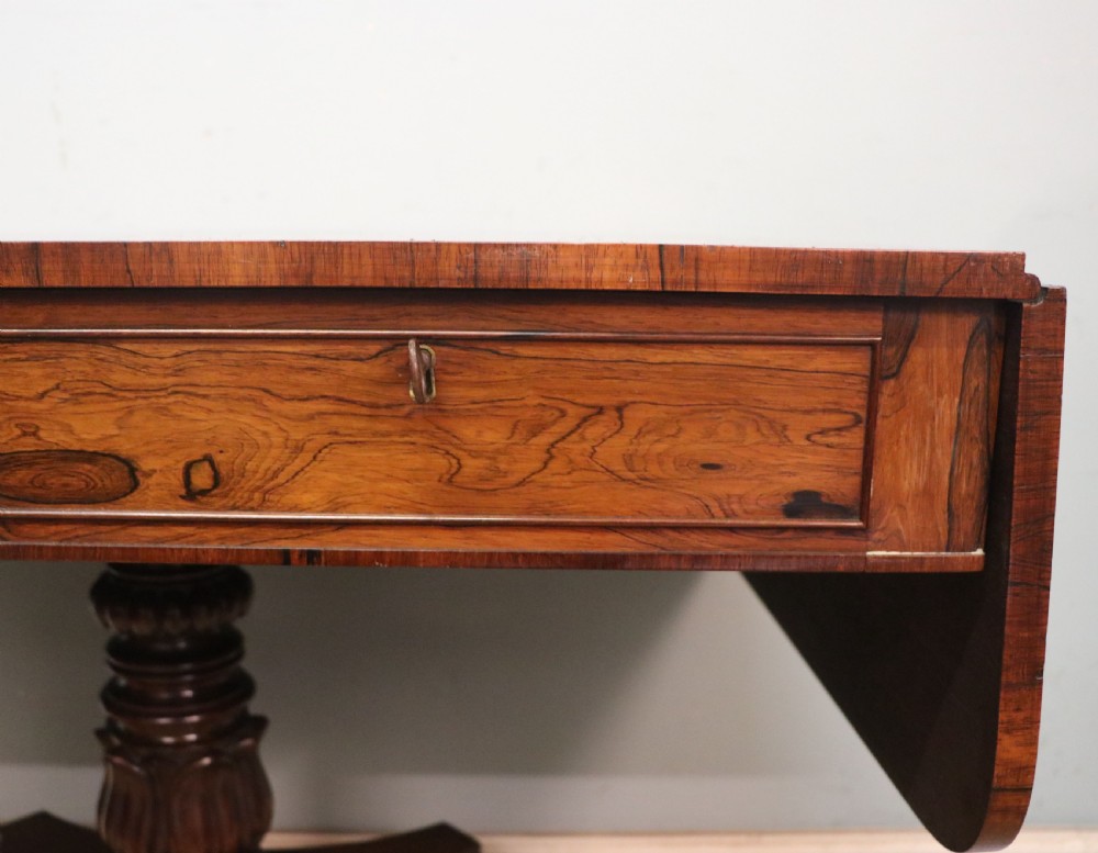 A late Georgian drop-leaf sofa table crafted from dark figured rosewood with a turned pedestal stem, two frieze drawers, and splayed quadruped legs with brass castors. View XV