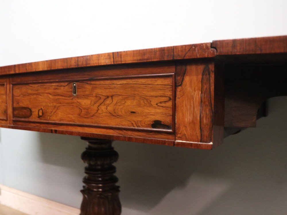 A late Georgian drop-leaf sofa table crafted from dark figured rosewood with a turned pedestal stem, two frieze drawers, and splayed quadruped legs with brass castors. View XVI