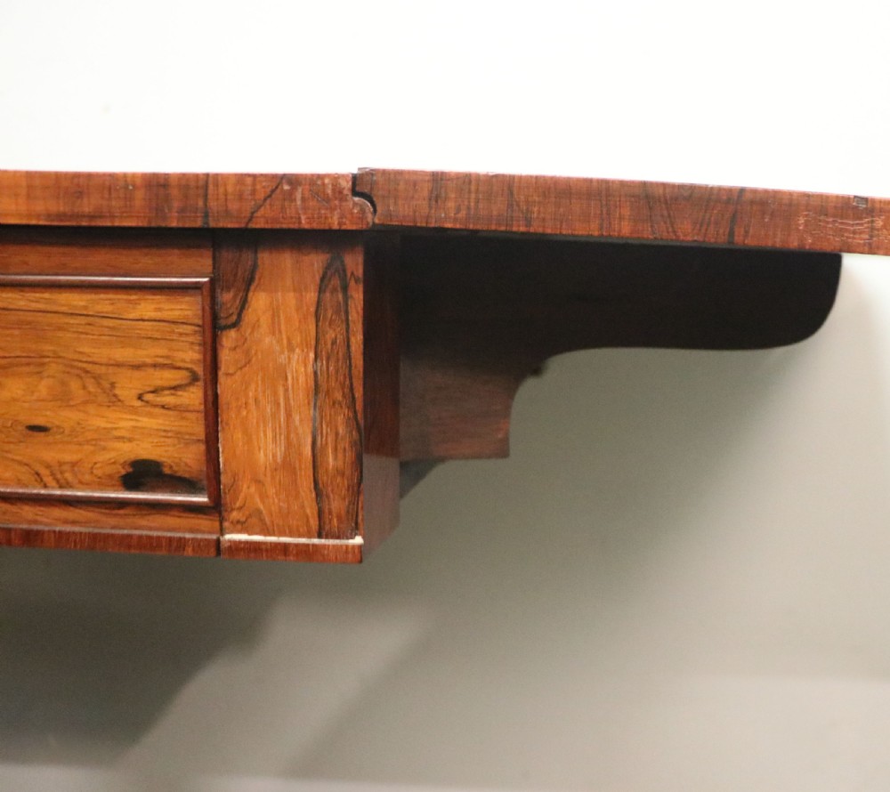 A late Georgian drop-leaf sofa table crafted from dark figured rosewood with a turned pedestal stem, two frieze drawers, and splayed quadruped legs with brass castors. View XVII