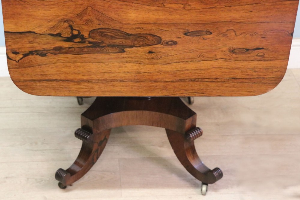 A late Georgian drop-leaf sofa table crafted from dark figured rosewood with a turned pedestal stem, two frieze drawers, and splayed quadruped legs with brass castors. View XXI