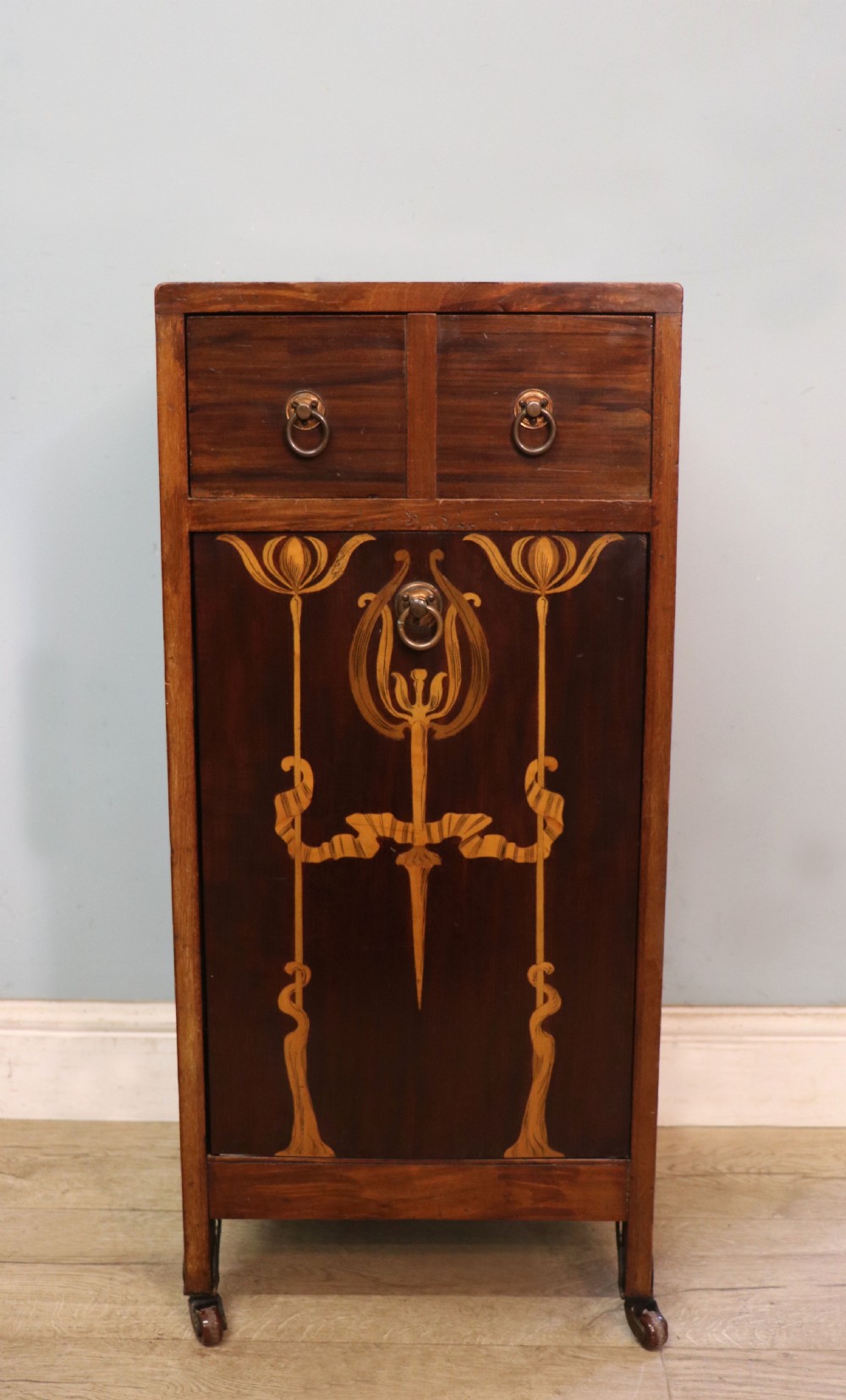 A tall dark wood Art Nouveau coal box featuring botanical marquetry inlay and brass ring handles, supported on castors.