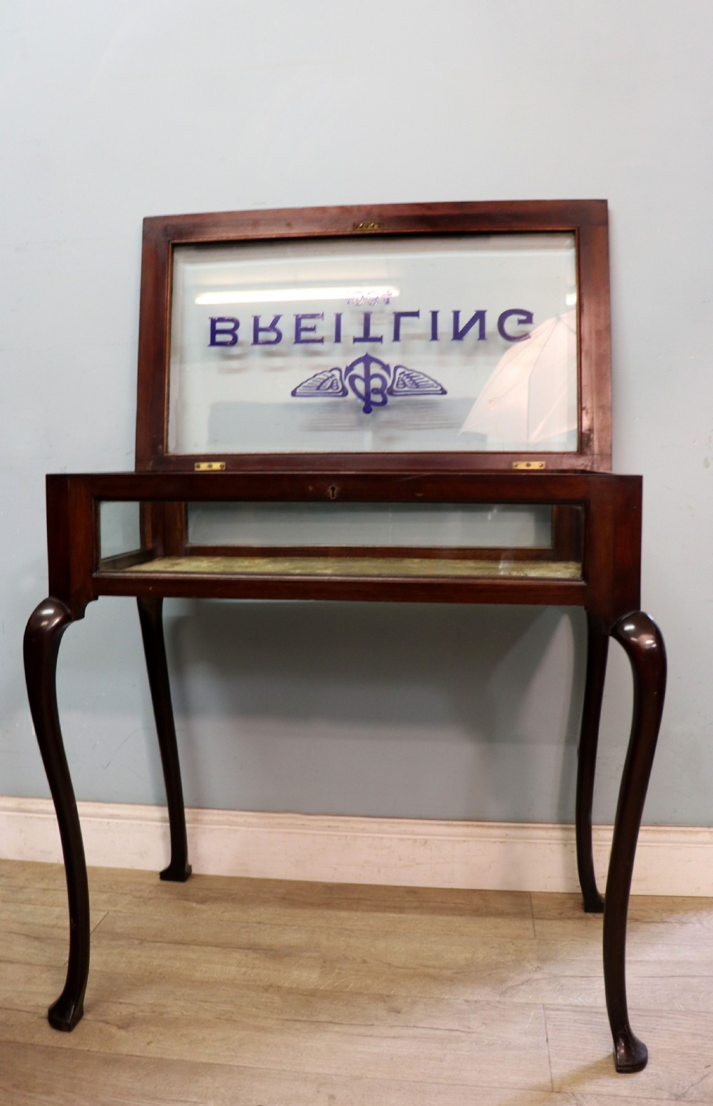 A dark mahogany glazed bijouterie table featuring cabriole legs, a glass top with blue Breitling watch branding, and a lined interior display compartment. View IV