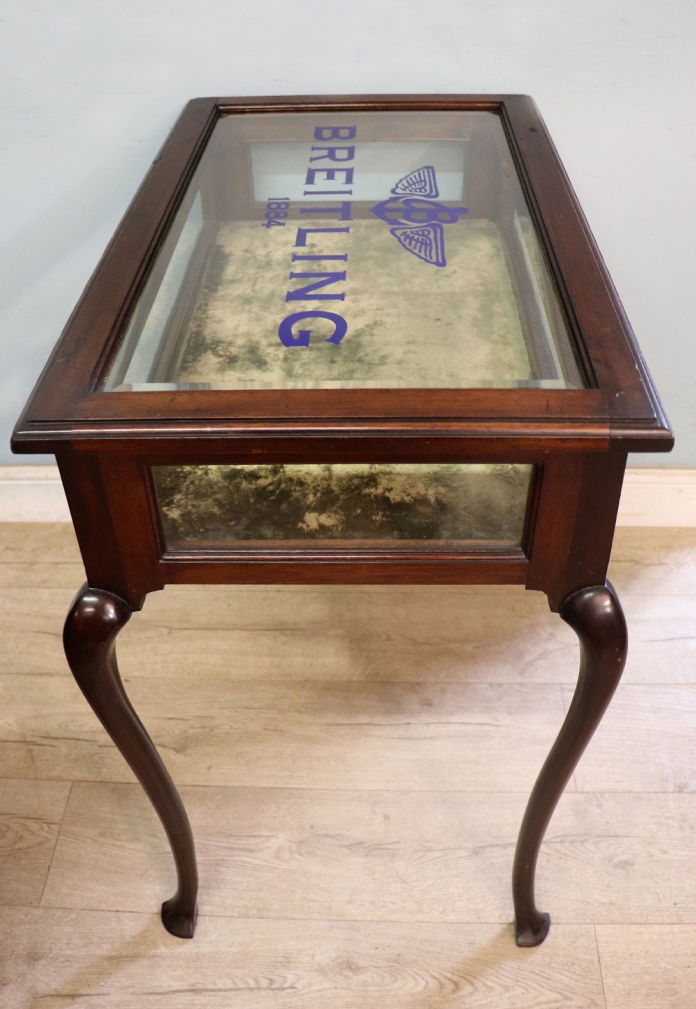 A dark mahogany glazed bijouterie table featuring cabriole legs, a glass top with blue Breitling watch branding, and a lined interior display compartment. View VI