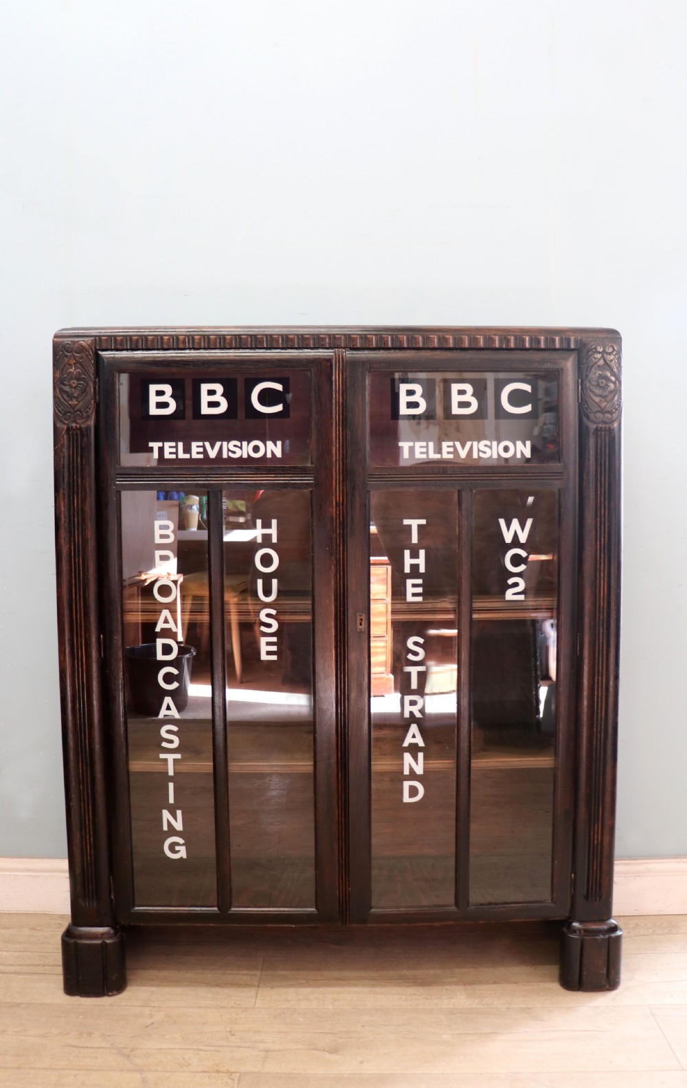 A dark oak glazed cabinet featuring hand-painted white lettering for the British Broadcasting Company across two glass doors.