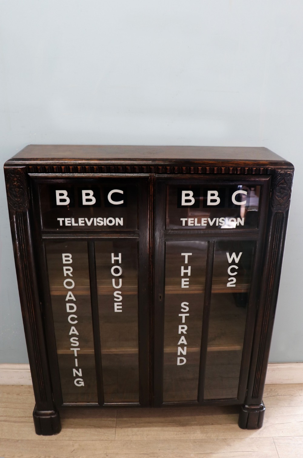 A dark oak glazed cabinet featuring hand-painted white lettering for the British Broadcasting Company across two glass doors. View II