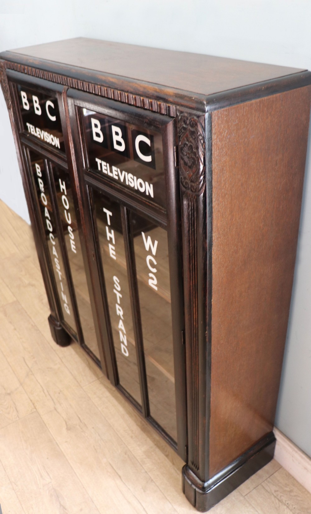 A dark oak glazed cabinet featuring hand-painted white lettering for the British Broadcasting Company across two glass doors. View IV
