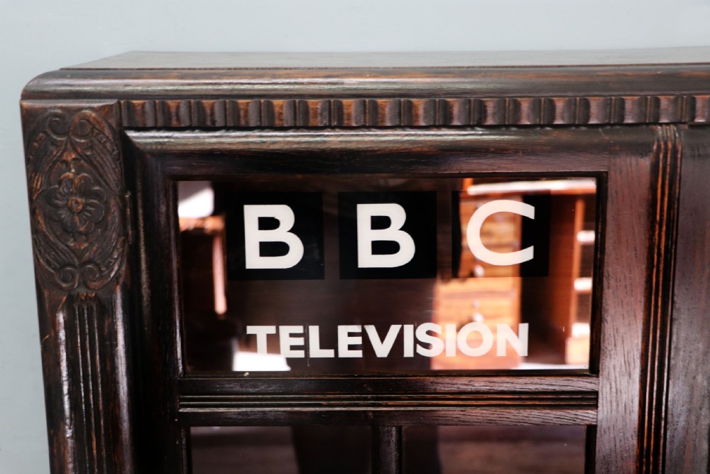 A dark oak glazed cabinet featuring hand-painted white lettering for the British Broadcasting Company across two glass doors. View V
