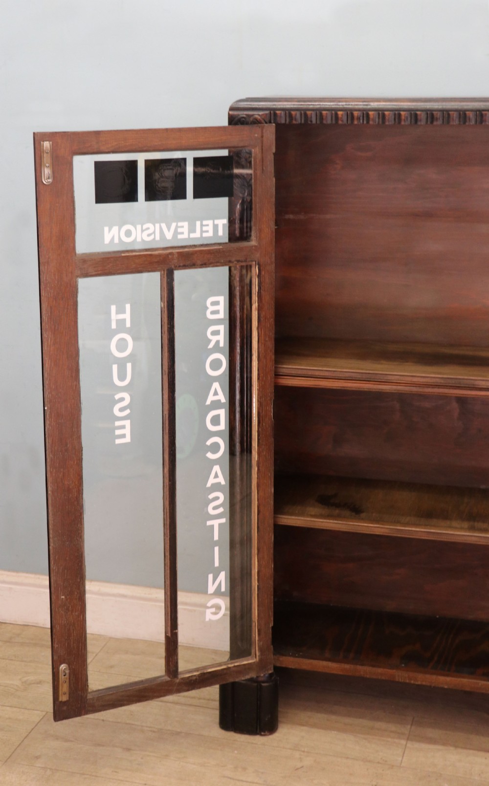 A dark oak glazed cabinet featuring hand-painted white lettering for the British Broadcasting Company across two glass doors. View IX