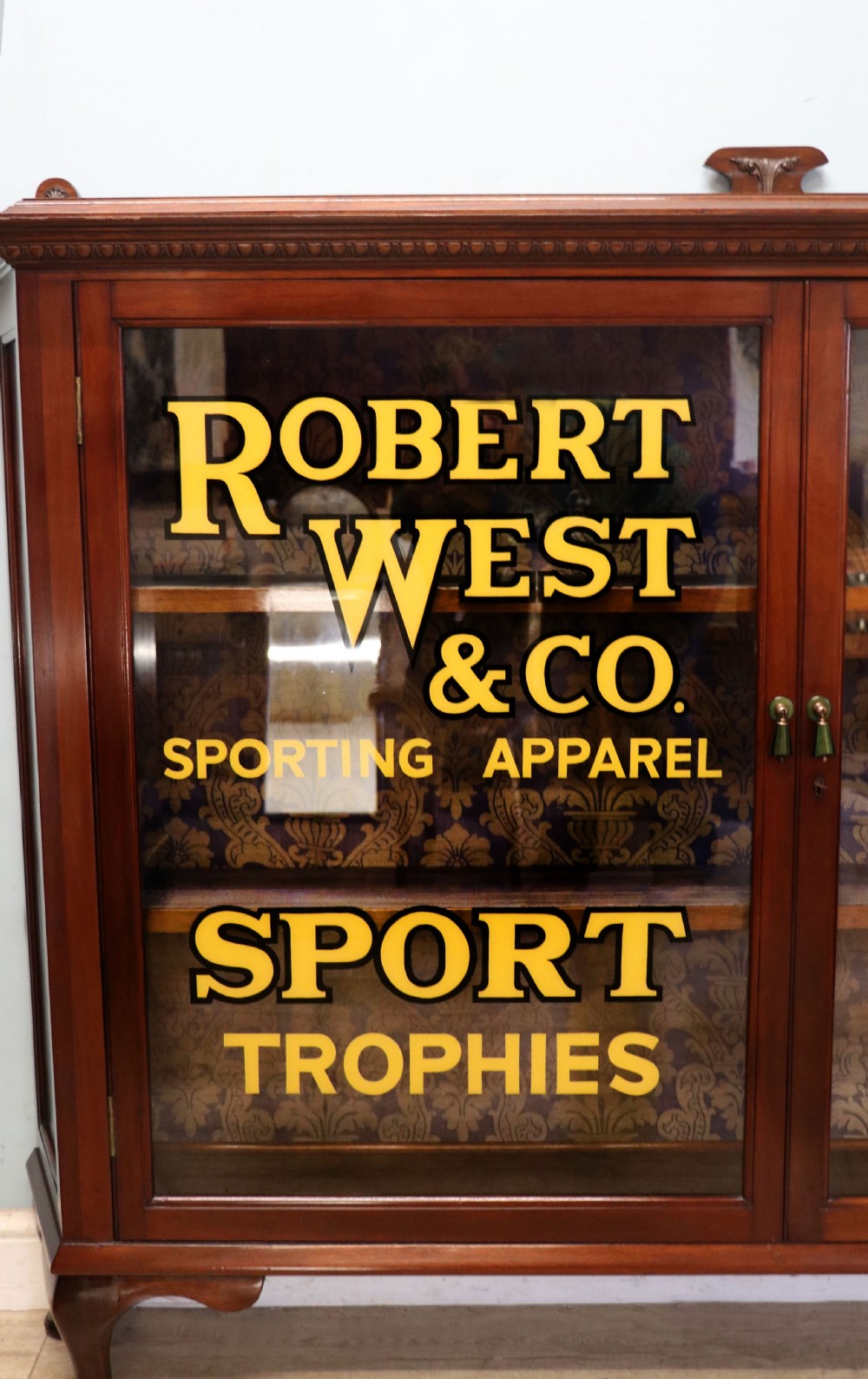 A late nineteenth-century dark mahogany display cabinet featuring two glazed doors adorned with hand-painted gold advertising typography, resting on short cabriole legs. View III