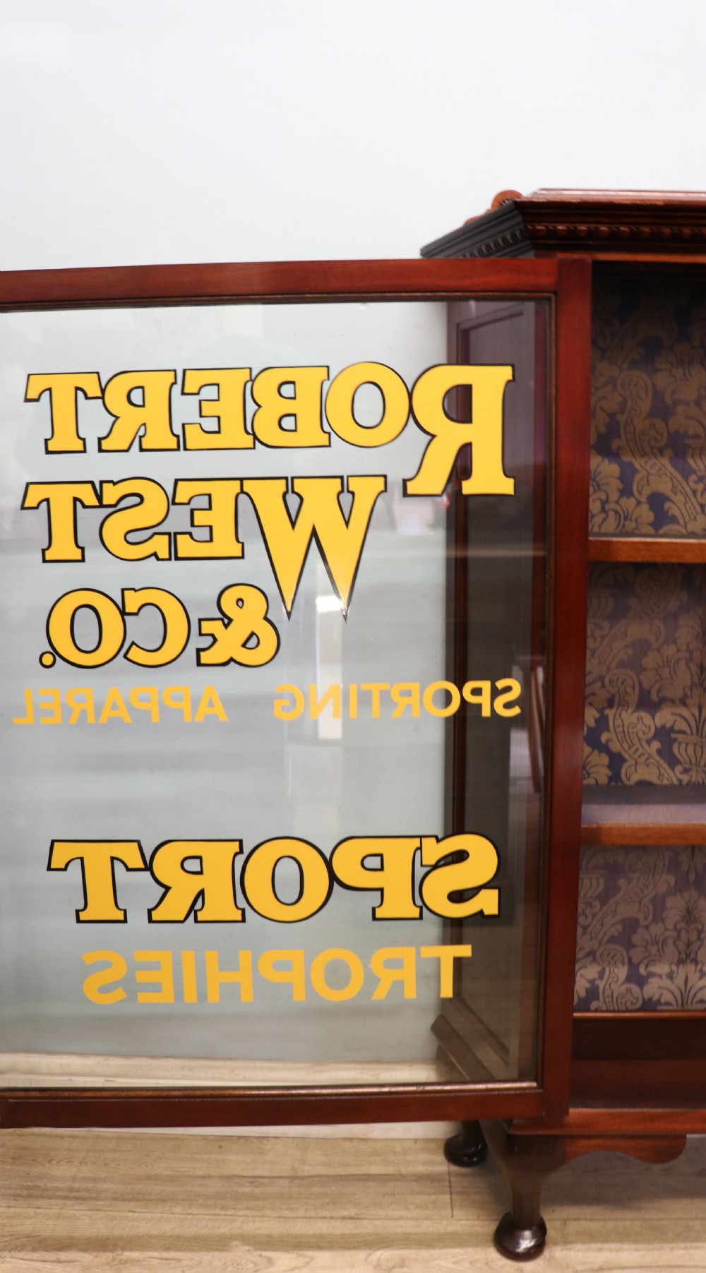 A late nineteenth-century dark mahogany display cabinet featuring two glazed doors adorned with hand-painted gold advertising typography, resting on short cabriole legs. View XIX